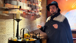 A person in historical attire, including a wide-brimmed hat and a cloak with lace detailing, holds a glass while standing beside a wooden table. The table has a plate with food items. Nearby, a wooden barrel is decorated with small pumpkins and orange candles. The background features rustic wall decor and cursive writing.