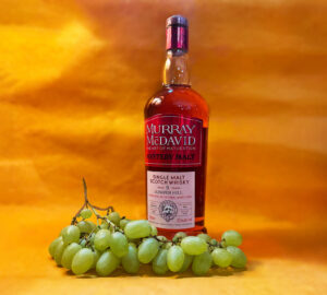 Bottle of Murray McDavid Juniper Hill 9 Scotch whisky agains a yellow/orange background accompanied with green grapes.