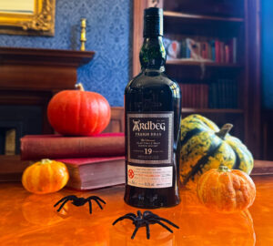 Bottle of Ardbeg 19 yer old Traigh Bhan Scotch whisky places on an amber table. There are two fake spiders places in front of the bottle. Around the bottle there are pumpkins in various colours and a stack of books to the left.