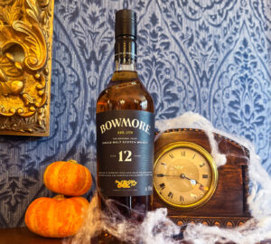 Bottle of Bowmore 12 year old Scotch whisky places on a wooden shelf with a old fashioned table clock covered with cobwebs. To the left of the bottle is 2 smll orange pumpkins.