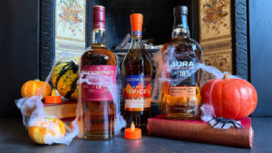 The image shows three bottles of Scotch whisky: The Glendronach Original 12, Glenmorangie Tale of Spices, and Jura 10. They are surrounded by small pumpkins, decorative squash, cobweb decorations, and a small orange candle. The background features a vintage fireplace with ornate tiles.
