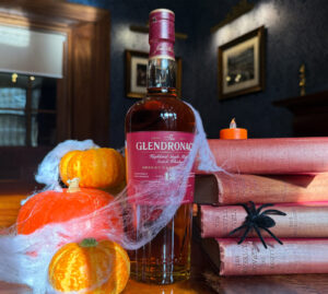 A bottle of Glendronach Scotch whisky is displayed on a wooden table. Surrounding it are small orange pumpkins, cobwebs, a black artificial spider, and stacked books with orange covers. A lit orange candle is also present, contributing to a Halloween-themed setup.