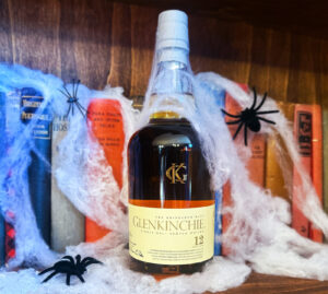 Bottle of Glenkinch 12 year old Scotch whisky placed on a wooden book shelf with old fashioned books. The bottle and books are adorned with cobwebs and spiders.