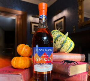 This image features a bottle of Glenmorangie "A Tale of Spices" Highland Single Malt Scotch whisky on a wooden table. Around it, there are a few decorative gourds, two orange pumpkins, and a small toy spider placed on old books. The background includes a dimly lit room with framed pictures on the walls.