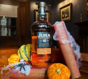 A bottle of Jura 10 Year Old Single Malt Scotch whisky is displayed on top of vintage Sherlock Holmes novels by Arthur Conan Doyle. Surrounding the bottle are small decorative pumpkins and a faux spider web with a plastic spider. The setting appears to be a warmly lit room with framed pictures on the walls.