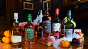 A selection of Scotch whisky bottles from brands such as Glenkinchie, Tomatin, Bowmore, Beinn Dubh, Isle of Skye, and Ardbeg are displayed on a wooden table. The scene is adorned with small pumpkins and faux cobwebs in a dimly lit room with wood paneling and framed art on the walls.