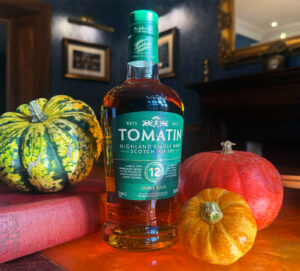 A bottle fo Tomatin 12 year old Scotch whisky is placed on a amber table with pumpkins in various colours.